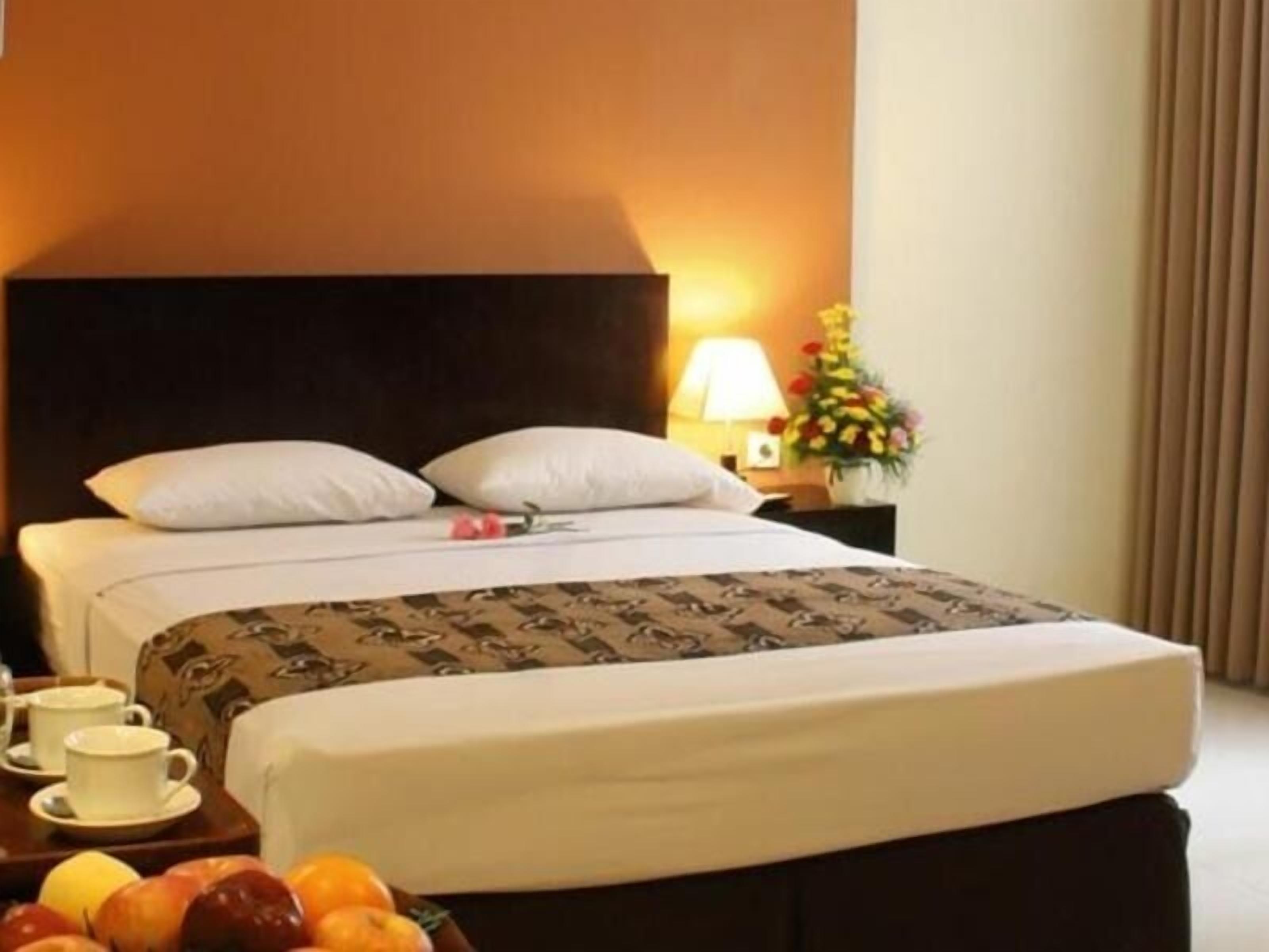 Una camera d'albergo con un letto, un comodino con una lampada, un vaso con fiori e un cesto di frutta.