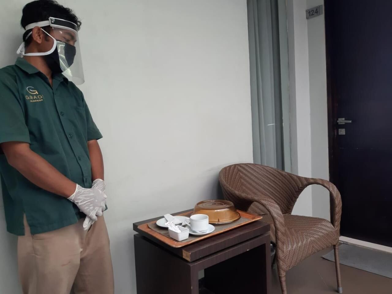 A hotel room with a wooden side table holding a tray with a teapot, cups, and a box. A wicker chair is against the wall.
