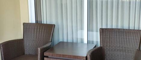 Un rincón con asientos que incluye dos sillas de mimbre y una pequeña mesa, una ventana grande con cortinas translúcidas y vistas a edificios.