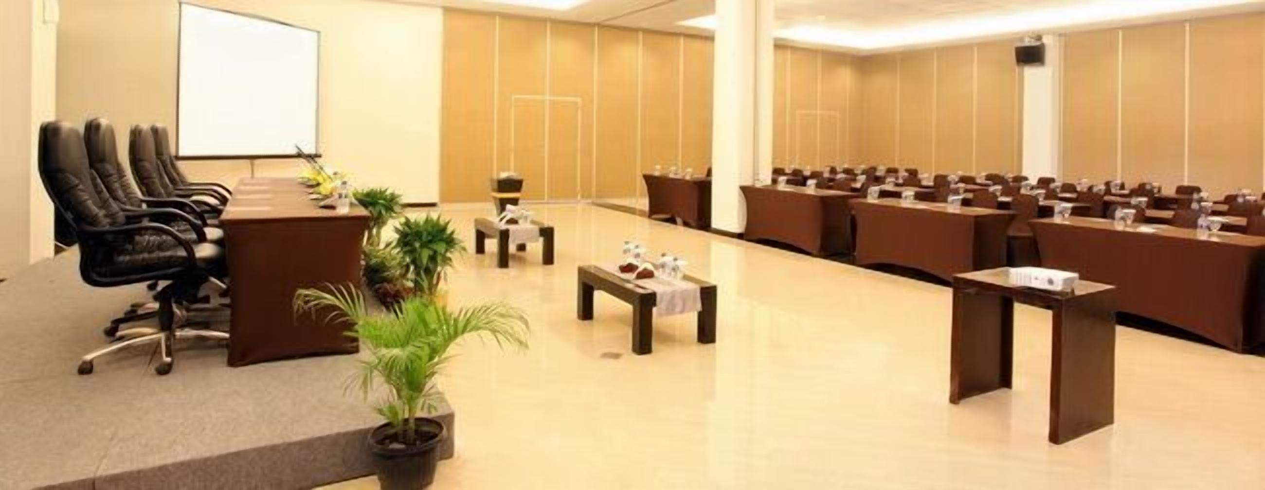 A conference room with a long table, multiple chairs, a projector screen, and a few potted plants.