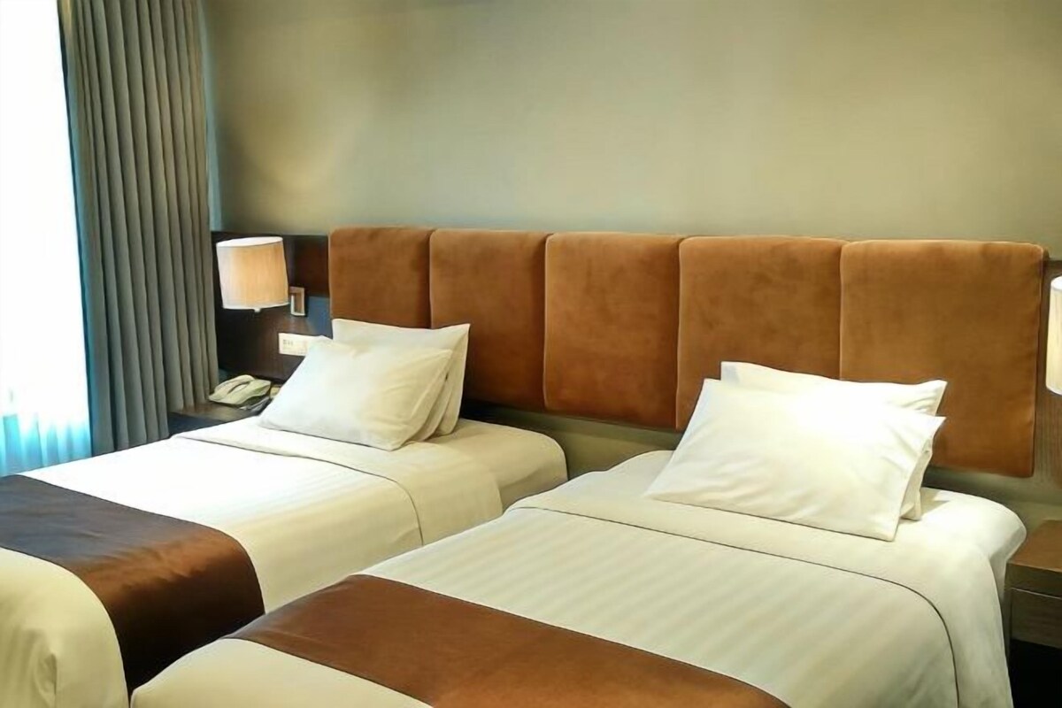 Ein Hotelzimmer mit zwei Betten, jeweils mit weißer Bettwäsche und braunen Kopfteilen. Auf der linken Seite befindet sich ein Nachttisch mit einer Lampe und einem Telefon.