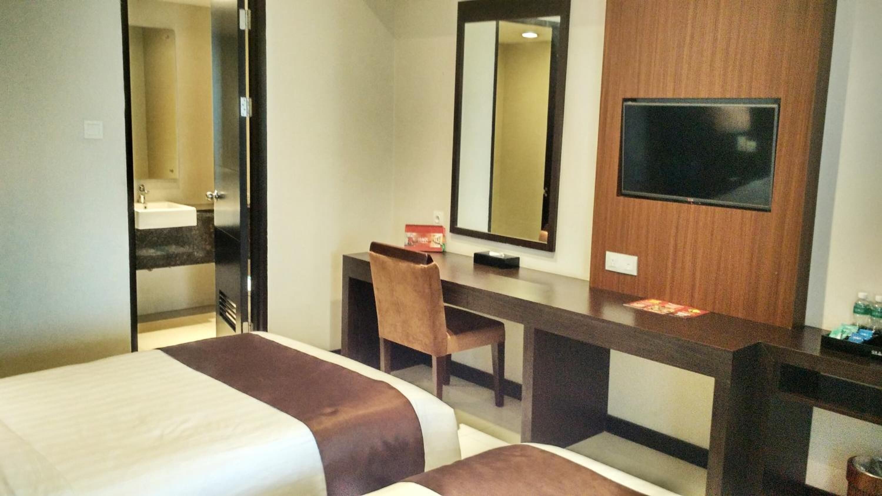 Habitación de hotel con dos camas, un escritorio, una silla, un espejo grande y un televisor en la pared.