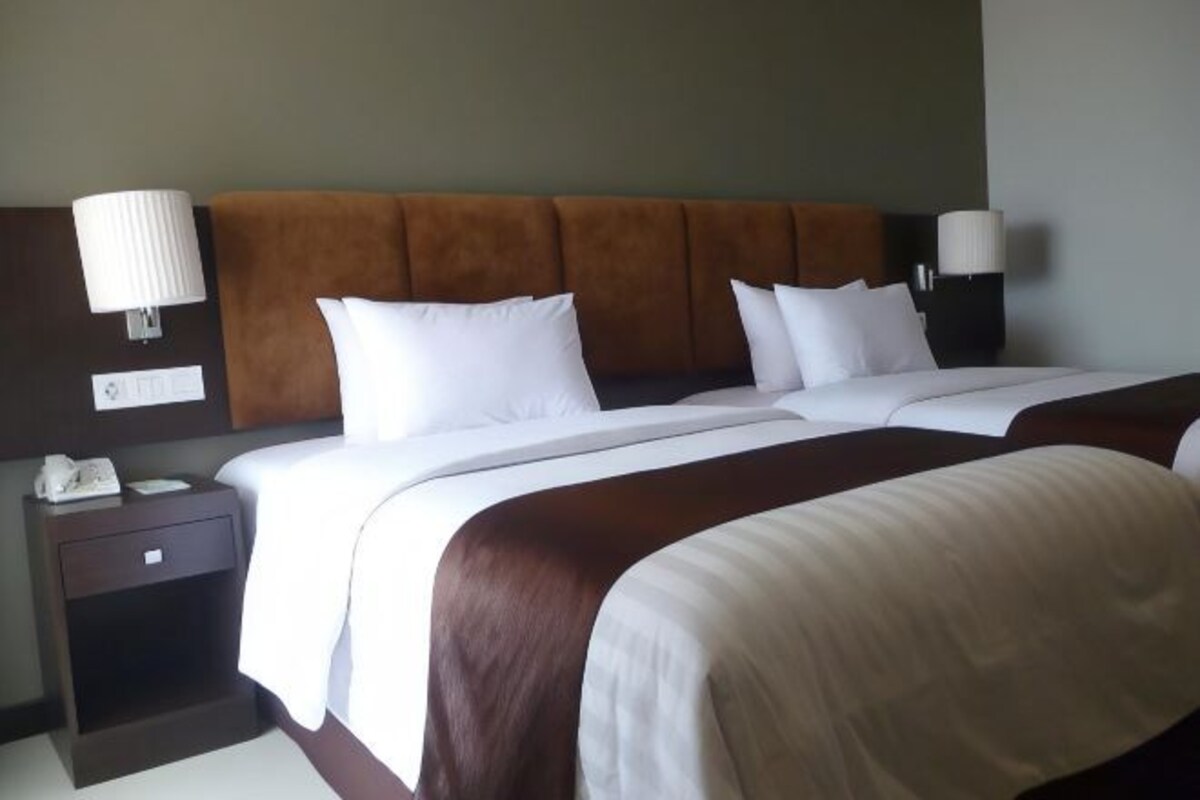 Een hotelkamer met twee bedden, een bruin hoofdeinde, wit beddengoed en bedlampjes.