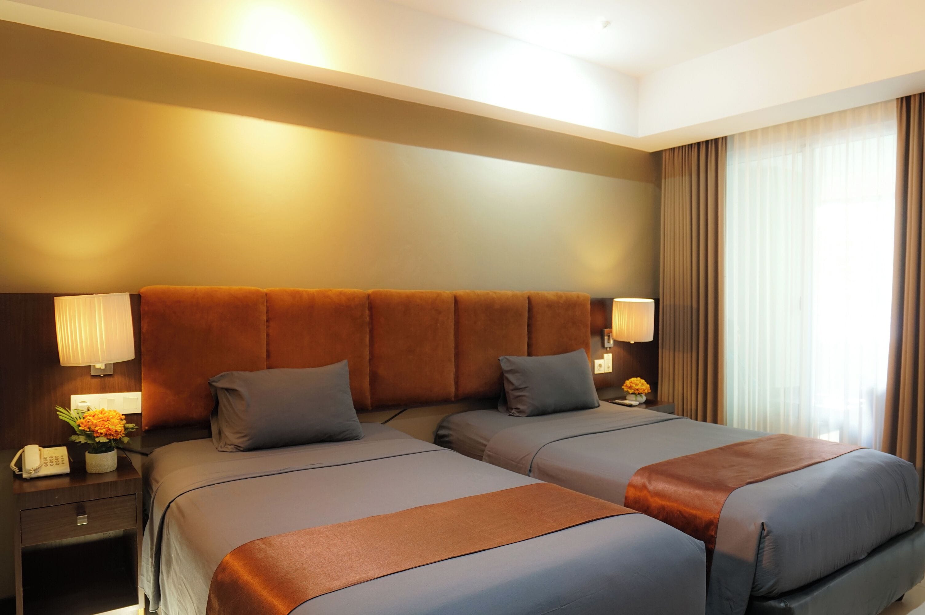 Une chambre d’hôtel avec deux lits, une tête de lit en bois, des lampes de chevet, une table de chevet avec un téléphone et une fenêtre avec des rideaux.
