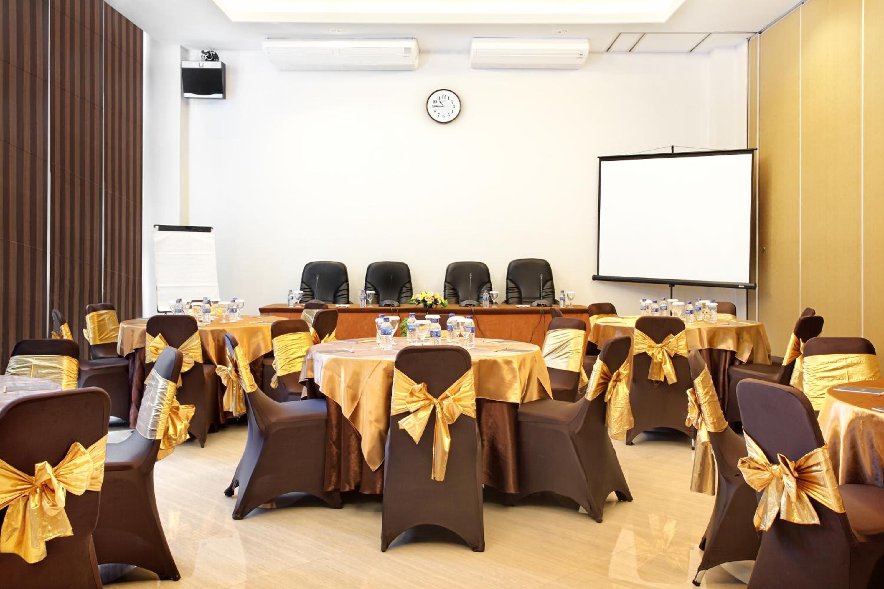Een vergaderzaal met ronde tafels, stoelen met gouden linten, een projectiescherm en een klok aan de muur.