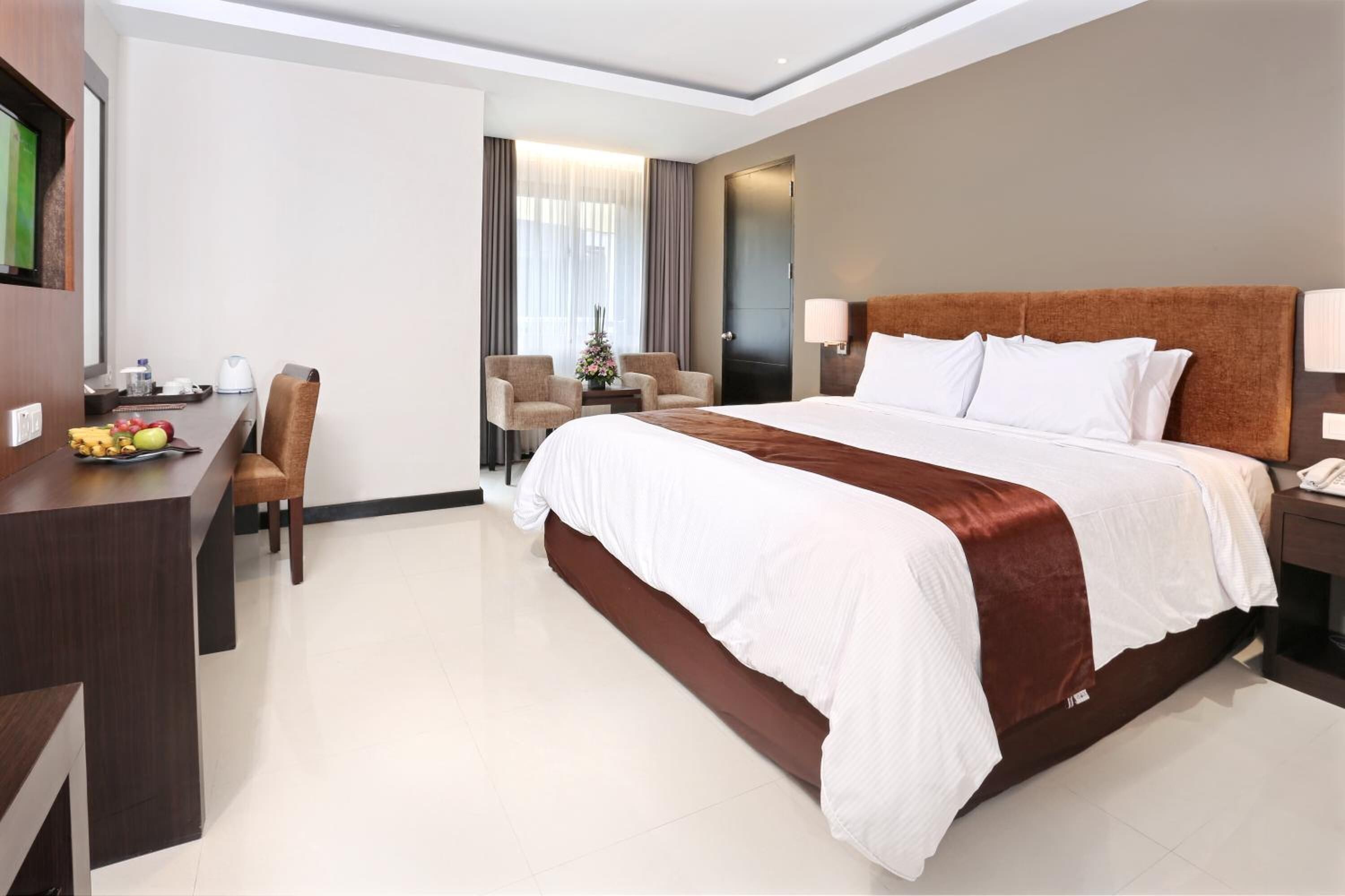 A hotel room with a large bed, a desk with a chair, a small table with a vase, and a window with curtains.