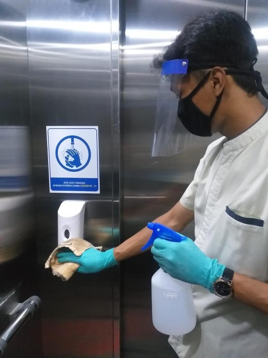 Una persona con camisa blanca y guantes azules limpiando una puerta de ascensor de acero inoxidable con un atomizador y un paño.