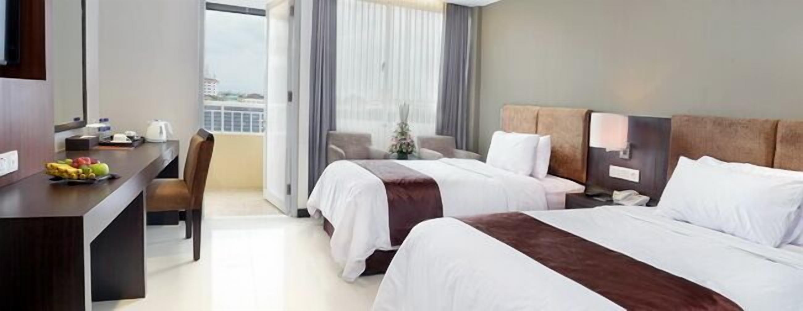 Habitación de hotel con dos camas, un escritorio con frutero, una silla y vista a la ciudad a través de amplias ventanas.
