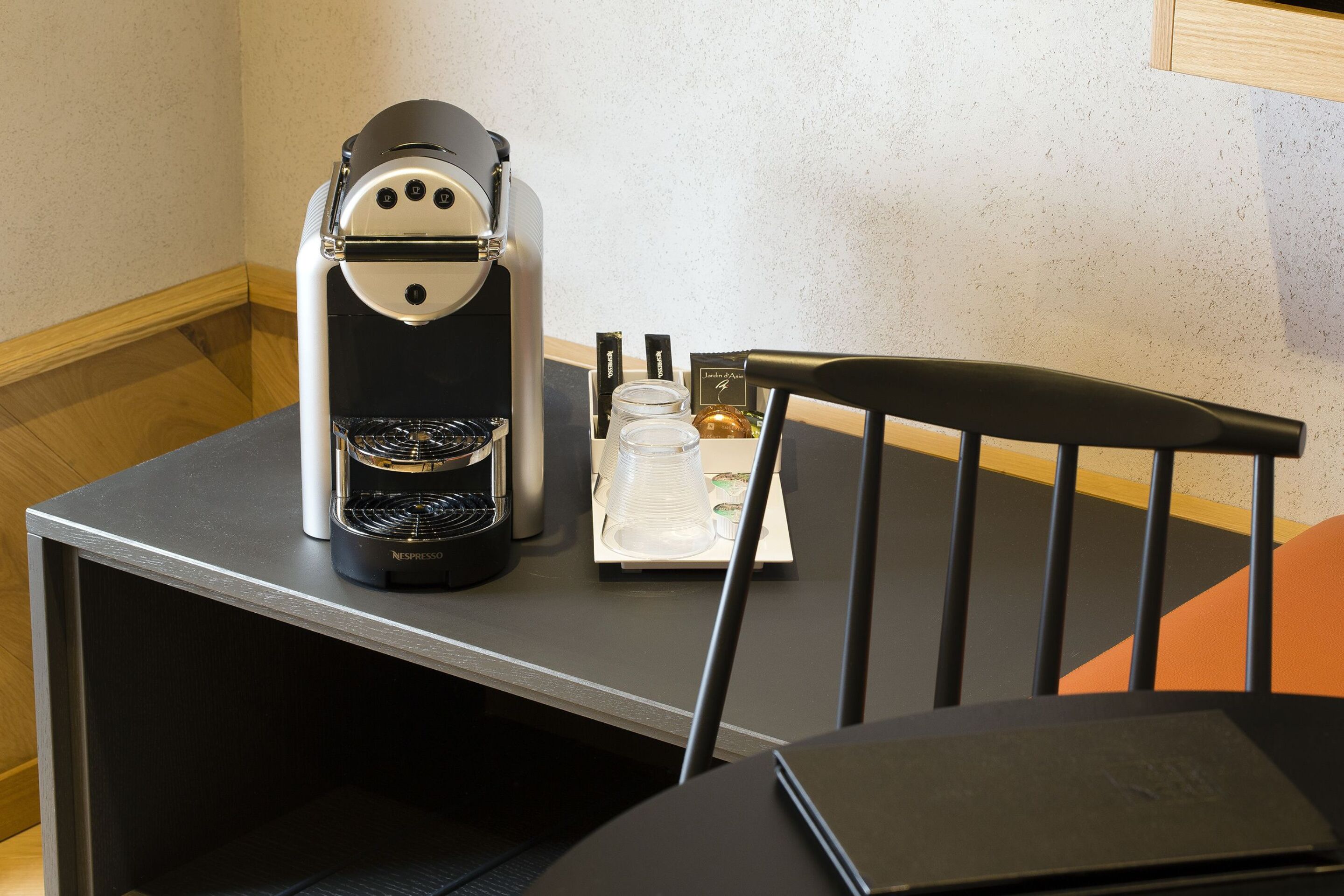 A Nespresso coffee machine on a table with a chair, a small table, and a bottle of hand sanitizer.