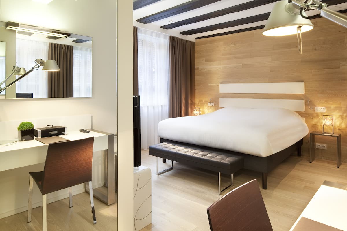 Ein modernes Hotelzimmer mit einem großen Bett, einem Schreibtisch mit Stuhl, einem Spiegel und einem Nachttisch mit Lampe.