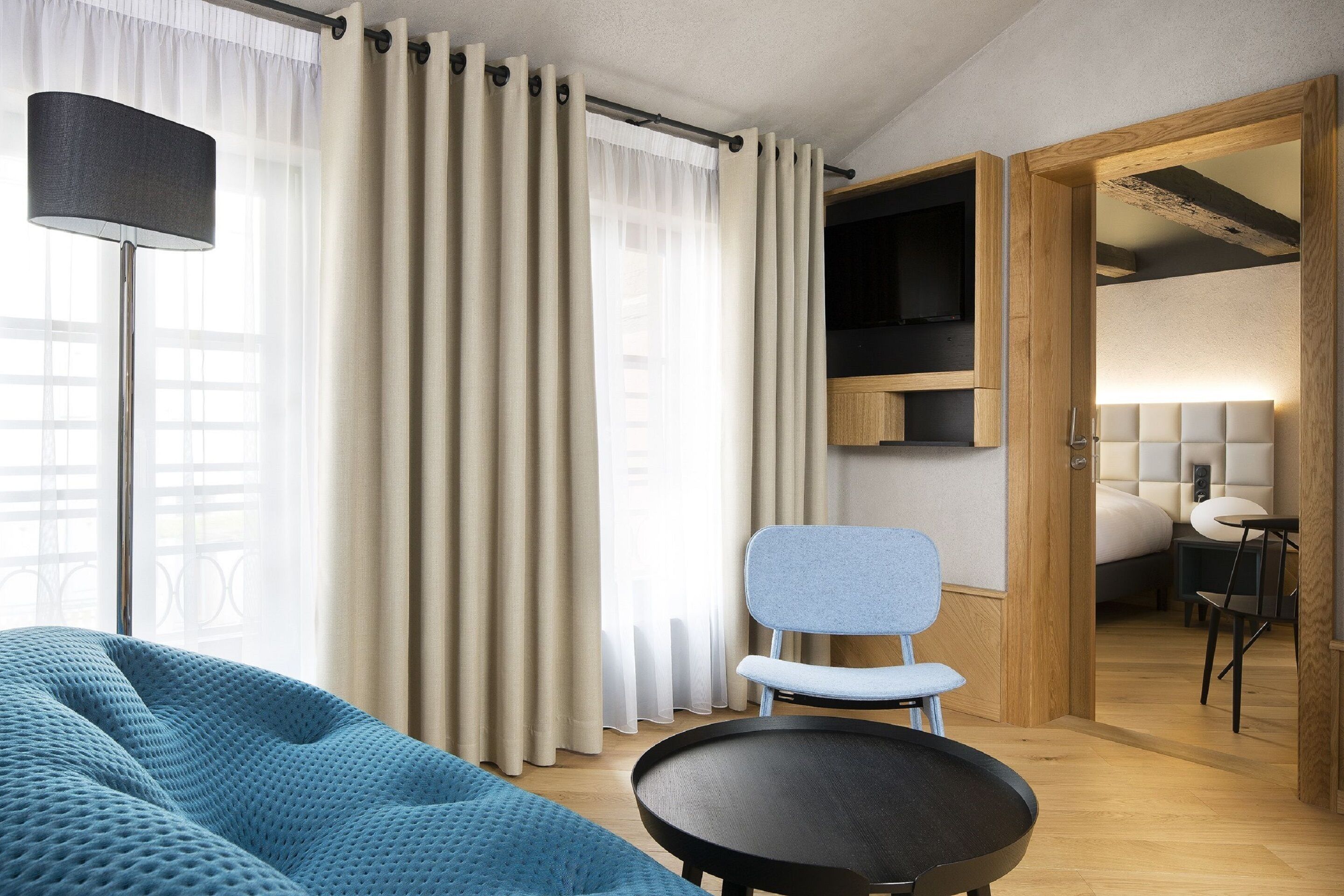 Una habitación de hotel moderna con una cama grande, un sofá azul, una mesa redonda y un televisor sobre una estantería de madera.