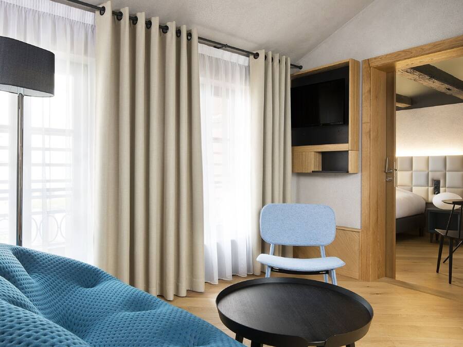 Una habitación de hotel moderna con una cama grande, un sofá azul, una mesa redonda y un televisor sobre una estantería de madera.