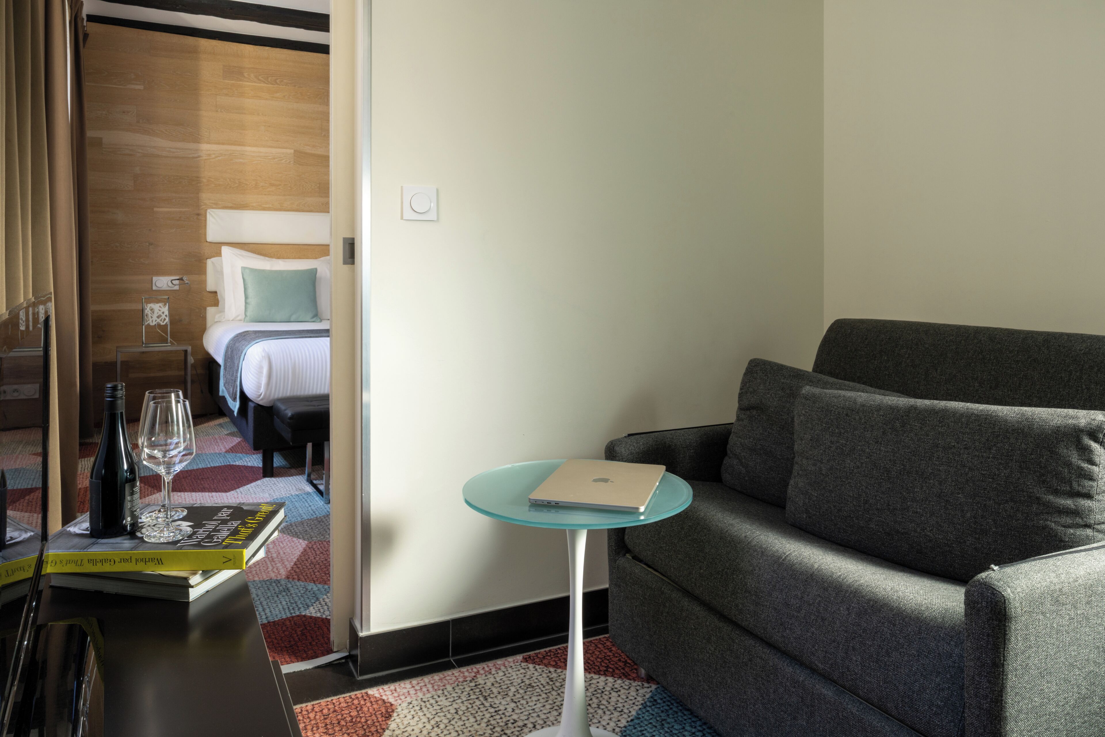 Una habitació d'hotel amb un llit, un sofà gris, una taula de vidre petita amb un portàtil, i una ampolla i un got decoratius sobre la taula auxiliar.