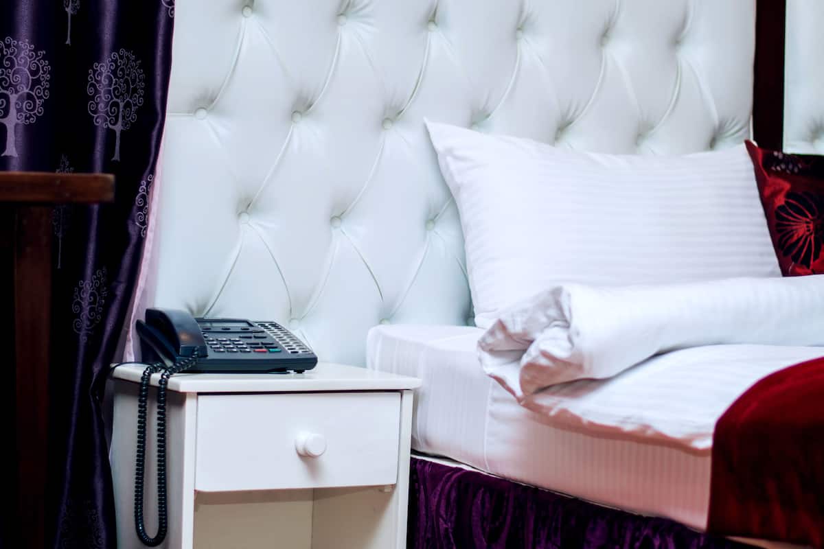 A neatly made bed with white bedding and red pillows, a bedside table with a telephone, and a dark curtain with a tree pattern.