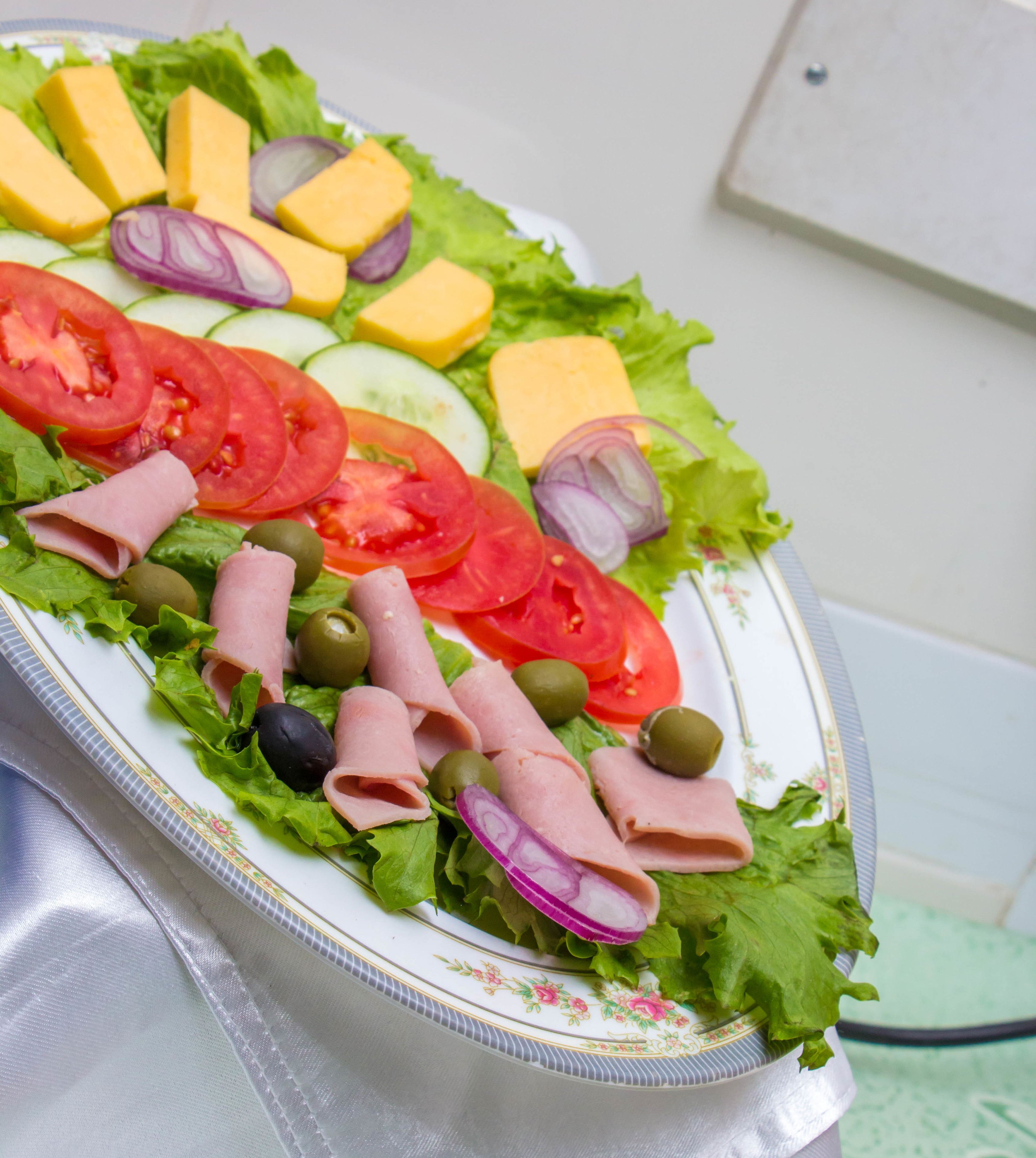 A plate of assorted deli items including ham, cheese, tomatoes, cucumbers, and olives on a decorative plate.