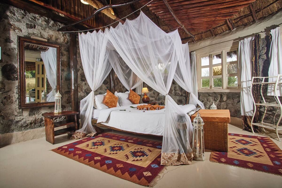 A room with a bed under a canopy, a nightstand, a lamp, a wicker chest, and a patterned rug.