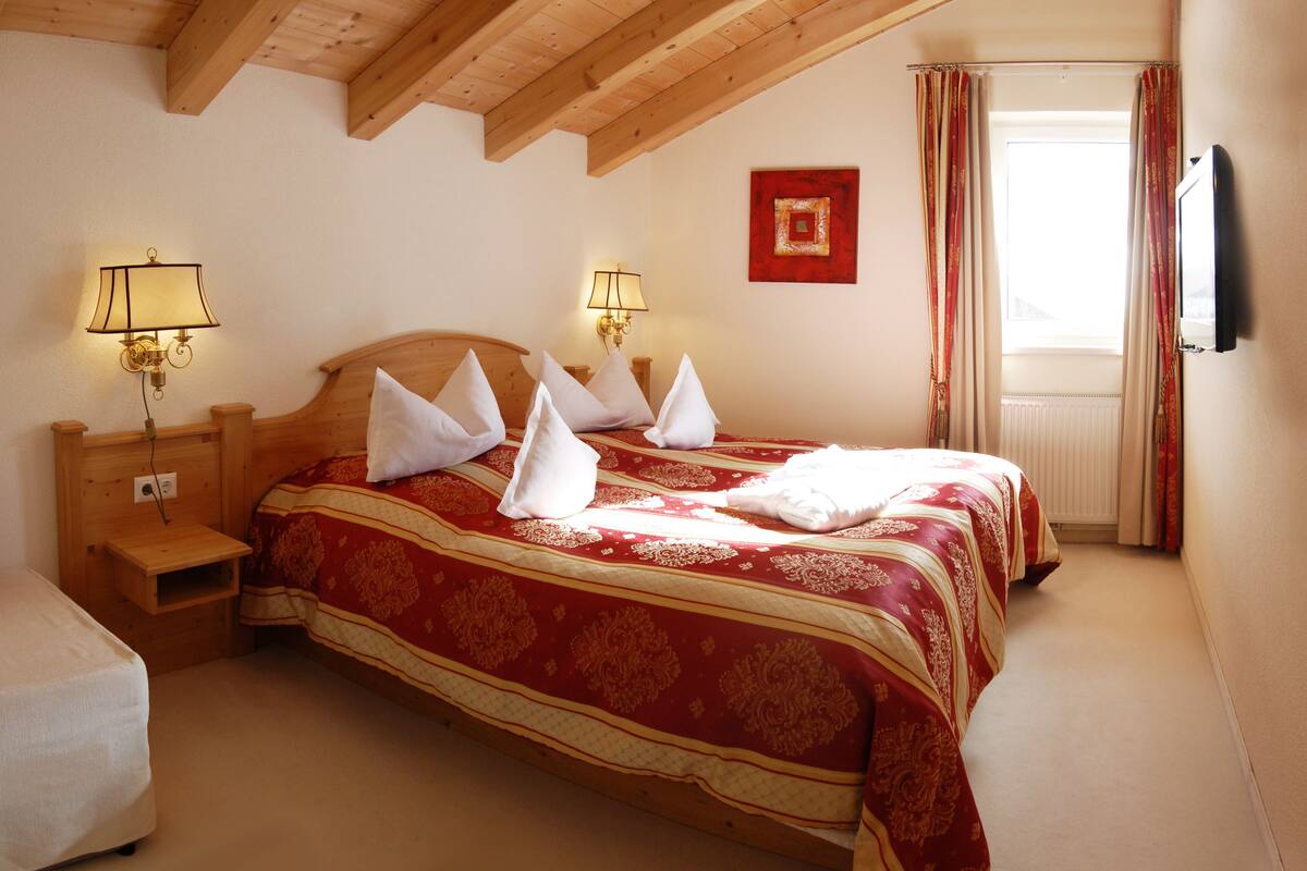 Un dormitorio con techo de madera, dos mesitas de noche con lámparas, una cama con ropa de cama roja y dorada, y una ventana con cortinas.