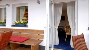 Een houten terras met een bank en stoelen, een plafondventilator en een deur die naar een binnenruimte leidt.