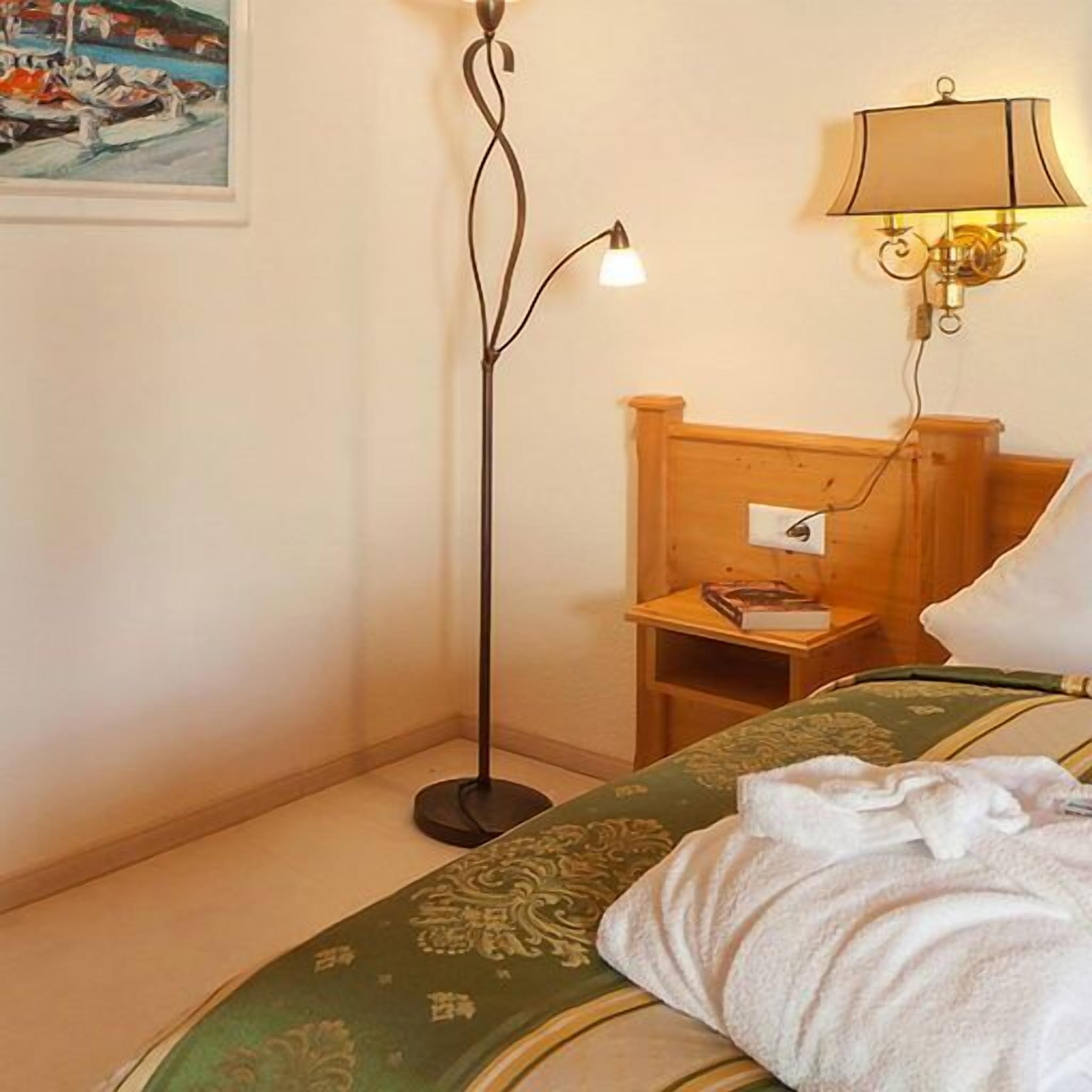 A hotel room with a bed, a floor lamp, a bedside table with a book, and a wall-mounted lamp.
