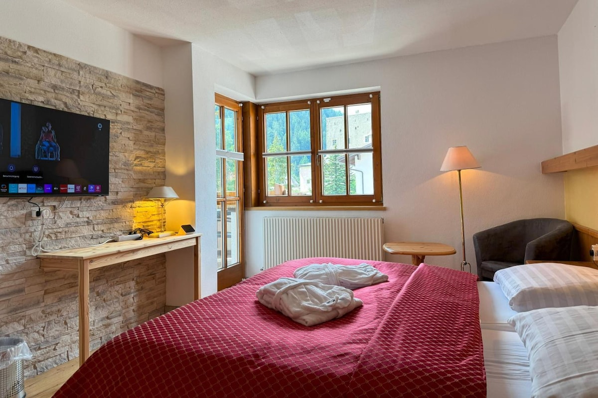 A hotel room with a bed, a desk with a computer, a TV mounted on the wall, a window, and a lamp.