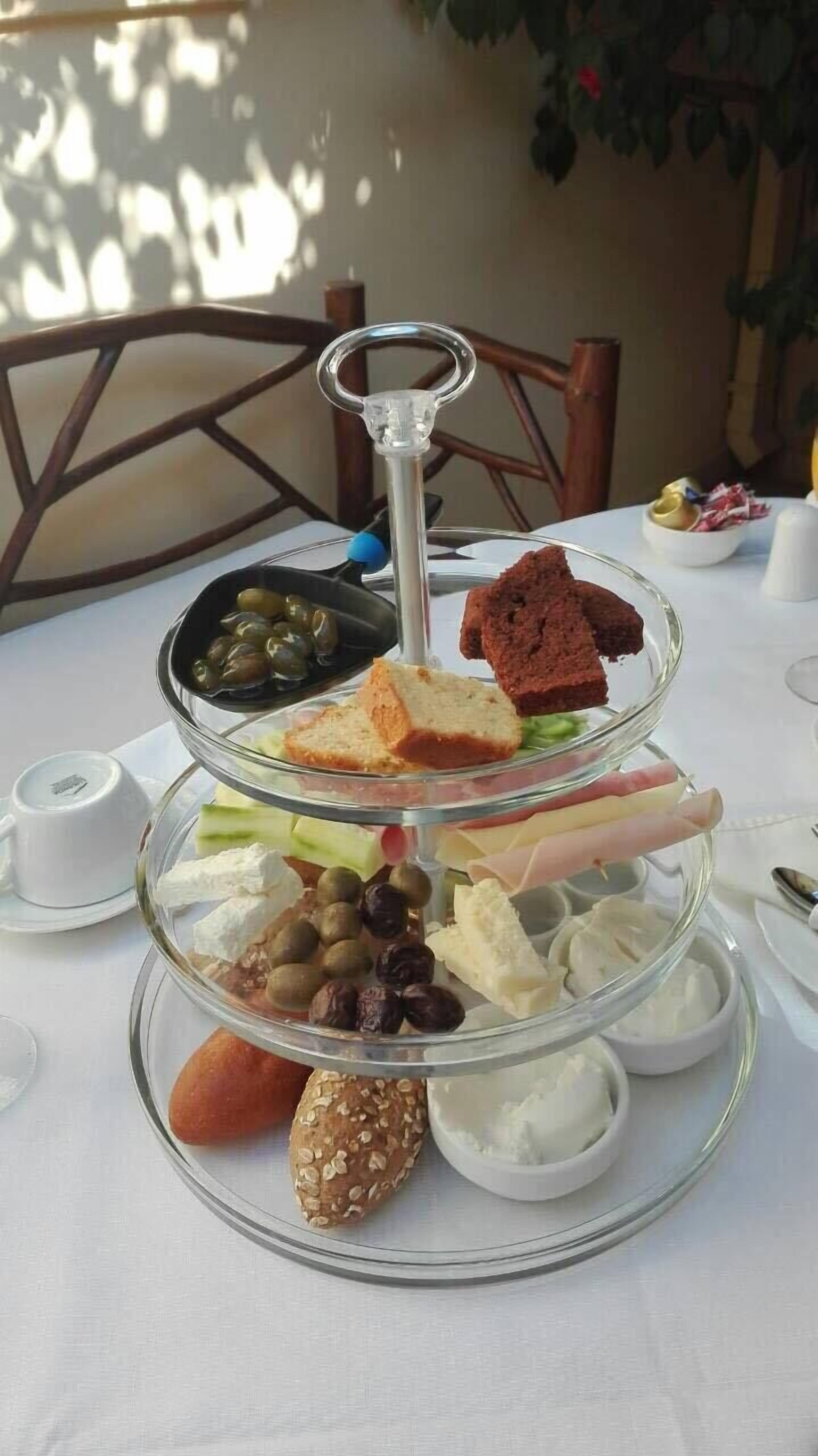 Ein mehrstufiges Glas-Serviertablett mit verschiedenen Speisen darunter Brot, Käse, Oliven und Nüsse.