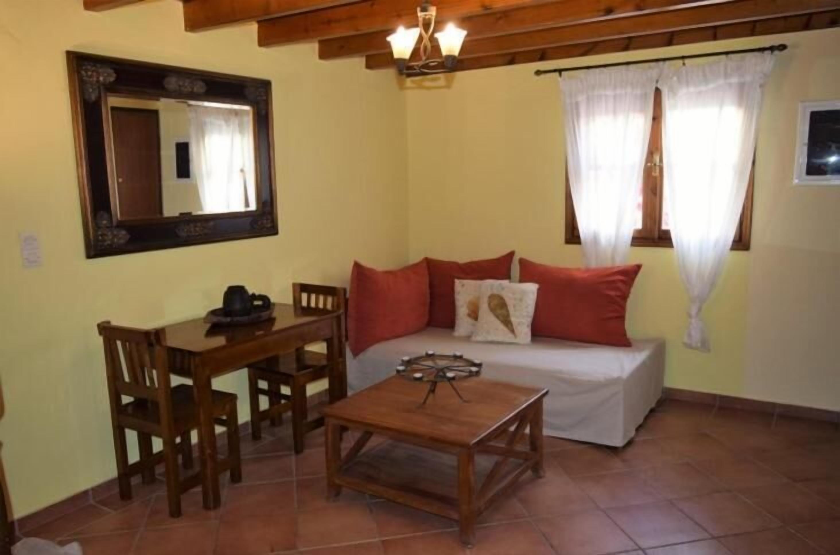 A cozy room with a wooden table, a sofa with red cushions, a mirror, and a window with curtains.