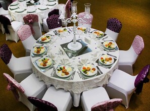 A round dining table with white chairs, each with a purple cushion. The table is set with plates of food, cutlery, and a central candelabra. - Hotel Adanava (Adana)