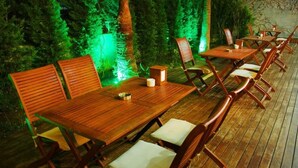 Een houten terras met tafels en stoelen, omringd door groen en verlicht met groene lampjes.