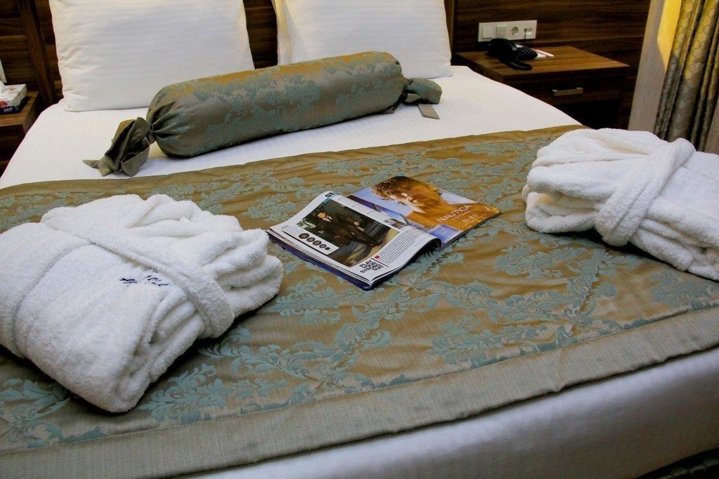 A neatly made bed with a magazine, two rolled towels, and a decorative pillow.
