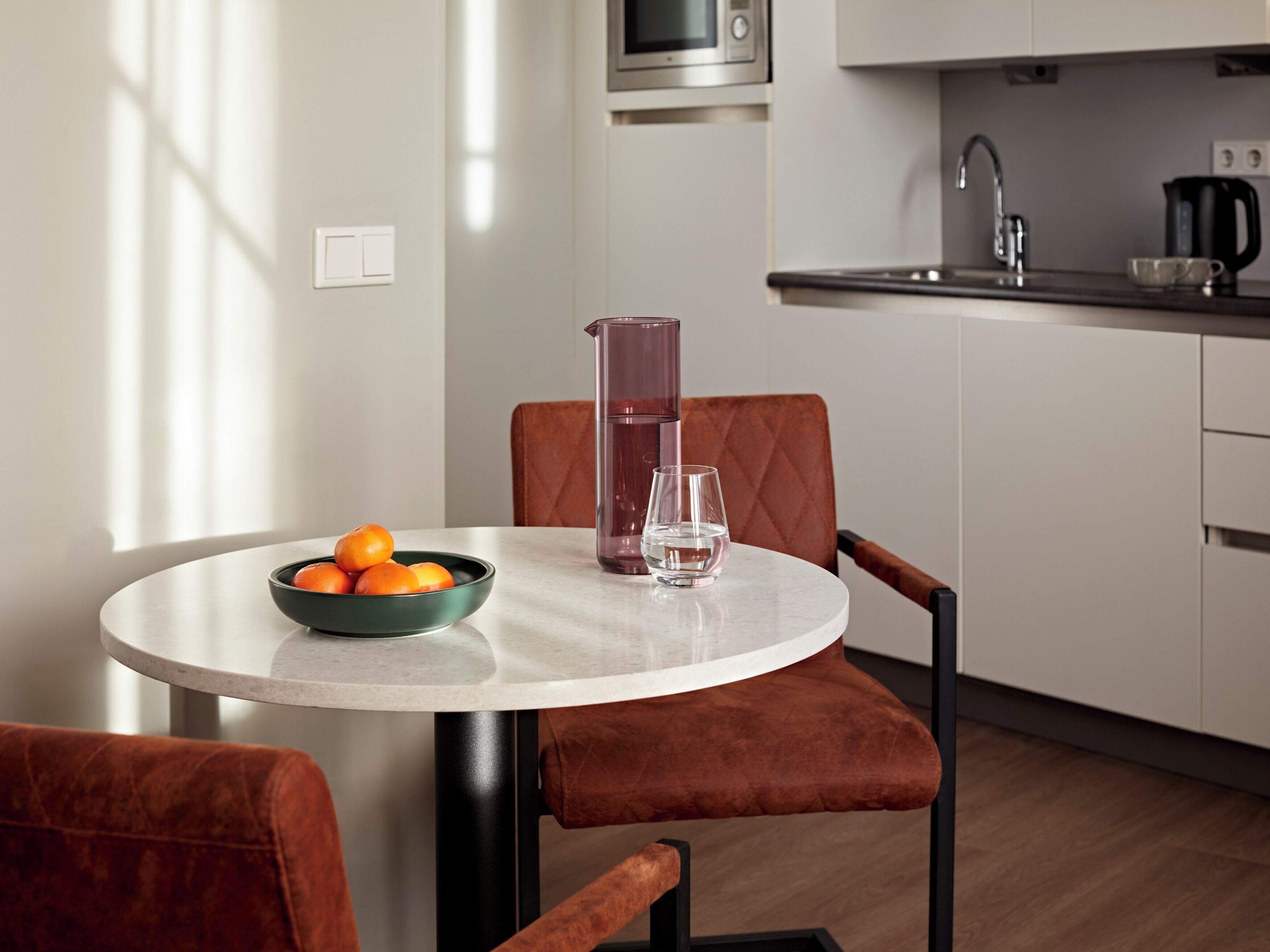 A modern kitchen with a round dining table, a bowl of oranges, a glass pitcher, and a glass of water.