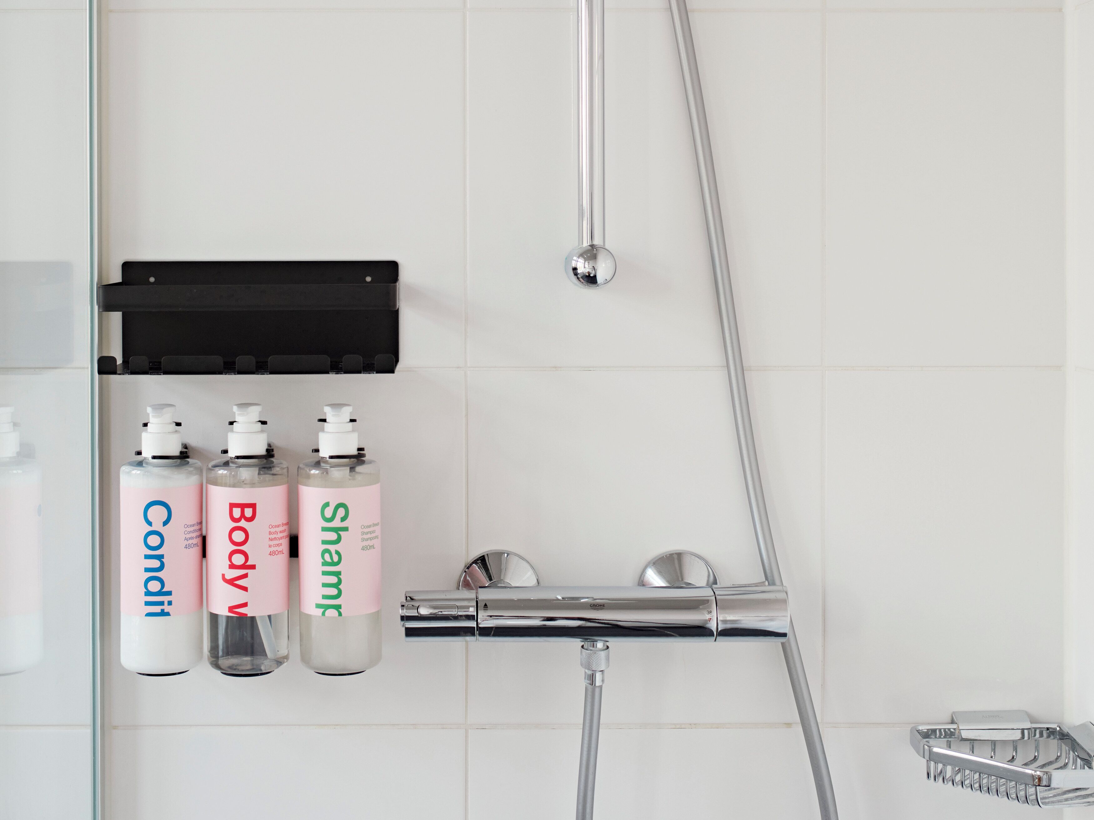 A bathroom with a glass shower enclosure, a chrome showerhead, and three bottles of body wash and shampoo on a shelf.