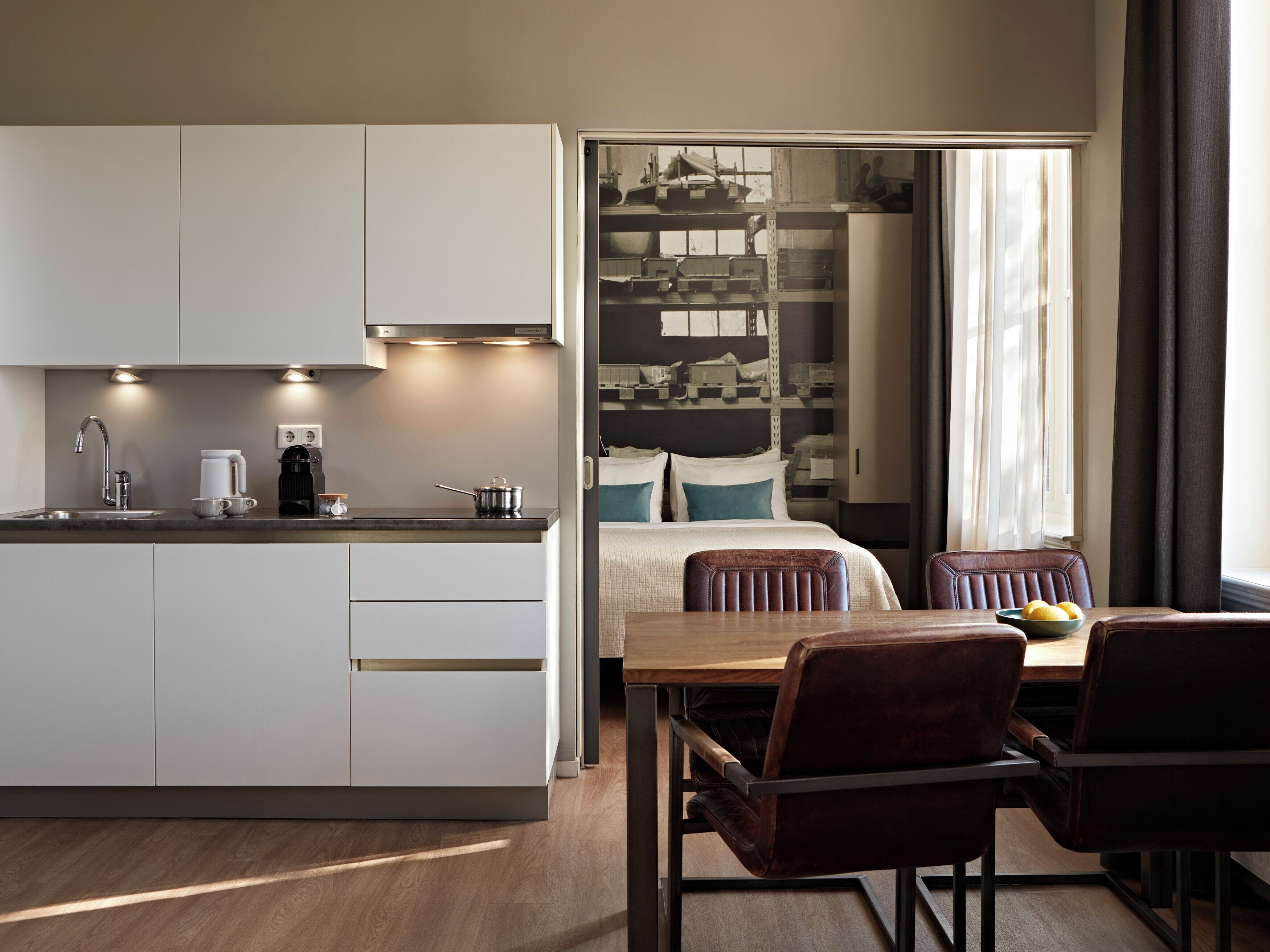 A modern kitchen with a dining area, a bed, and a view of a cityscape through a window.