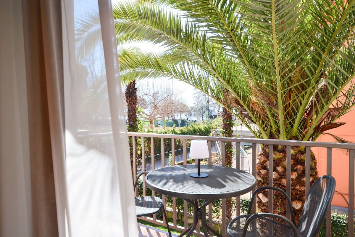 A balcony with a round table and chairs, a lamp, and a view of palm trees and a building in the background.