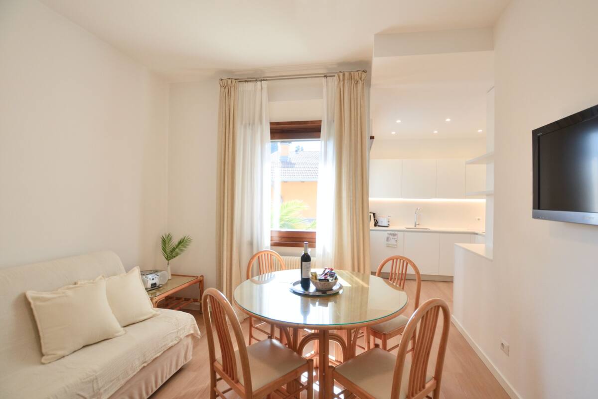 A modern living room with a glass dining table, beige sofa, and wooden chairs. A TV is mounted on the wall, and a window with curtains is visible.