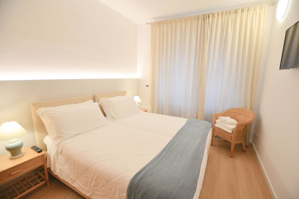 A neatly made bed with white linens, a bedside lamp, and a wicker chair with a towel.
