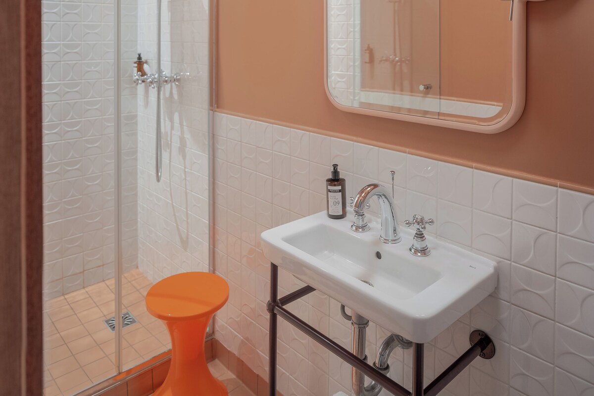 A bathroom with a glass-enclosed shower, a white sink with a modern faucet, and a round mirror with two lights.