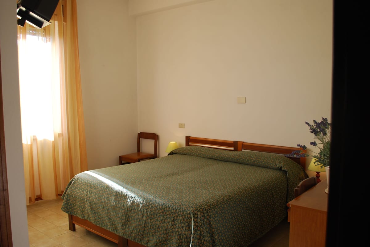 Une chambre à coucher avec un lit, une chaise, une table de chevet, une lampe et une fenêtre avec des rideaux.