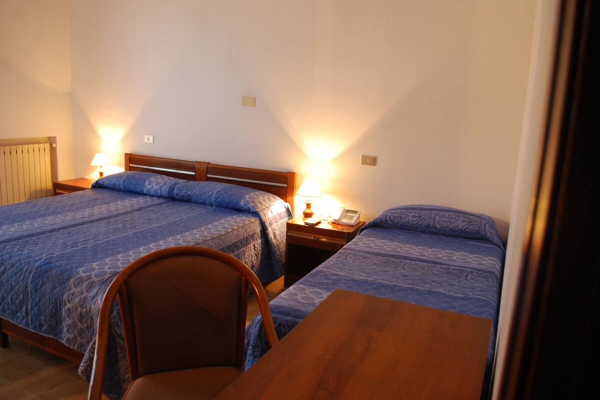 Une chambre avec deux lits, chacun doté d’une tête de lit en bois, d’une table en bois et d’une chaise. Chaque lit est muni d’une lampe de chevet.