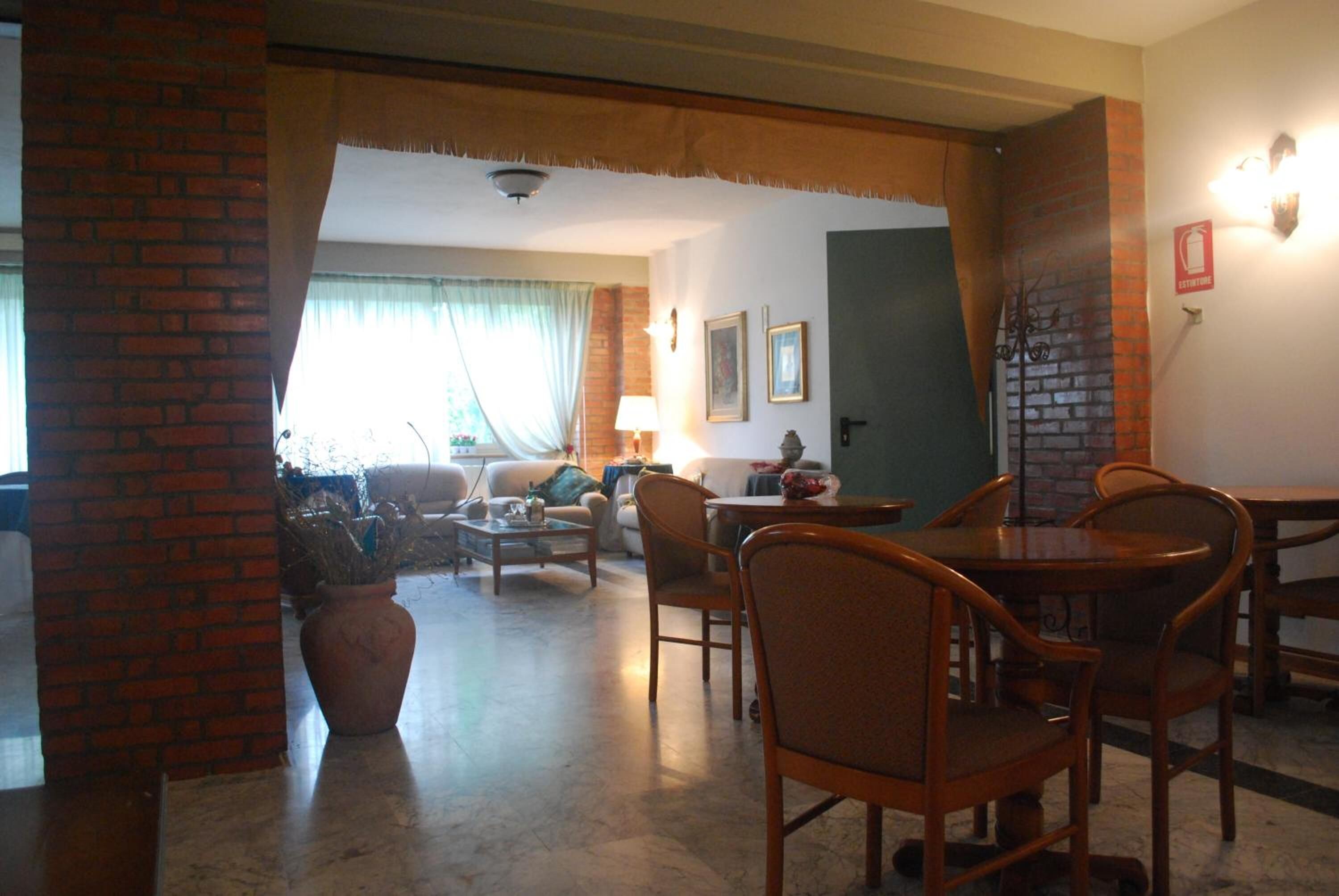 A living room with a brick column, a dining area with wooden chairs, a table, and a sofa. There is a potted plant and a lamp on a side table.