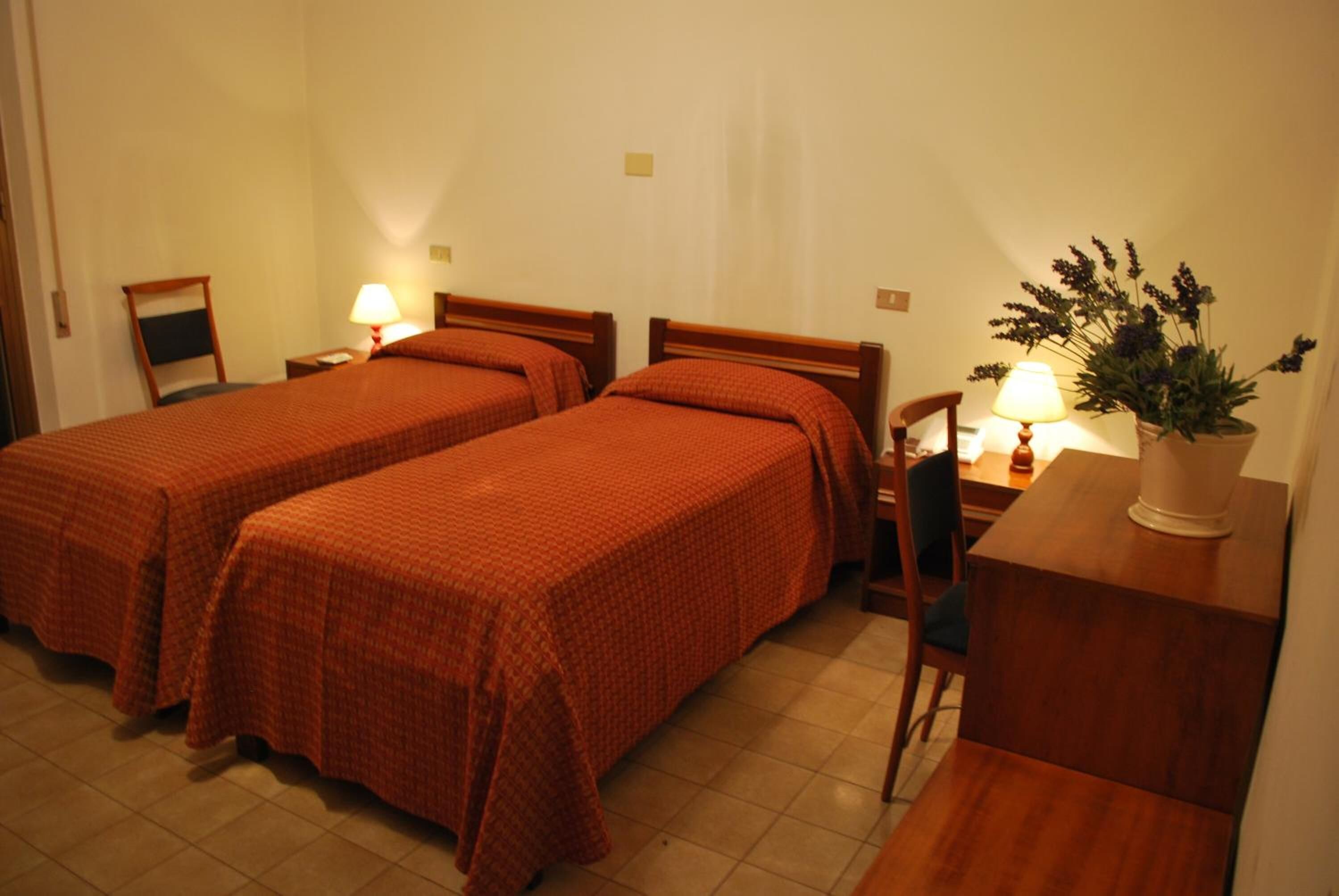 A hotel room with two single beds, a desk, a chair, a lamp, and a potted plant.