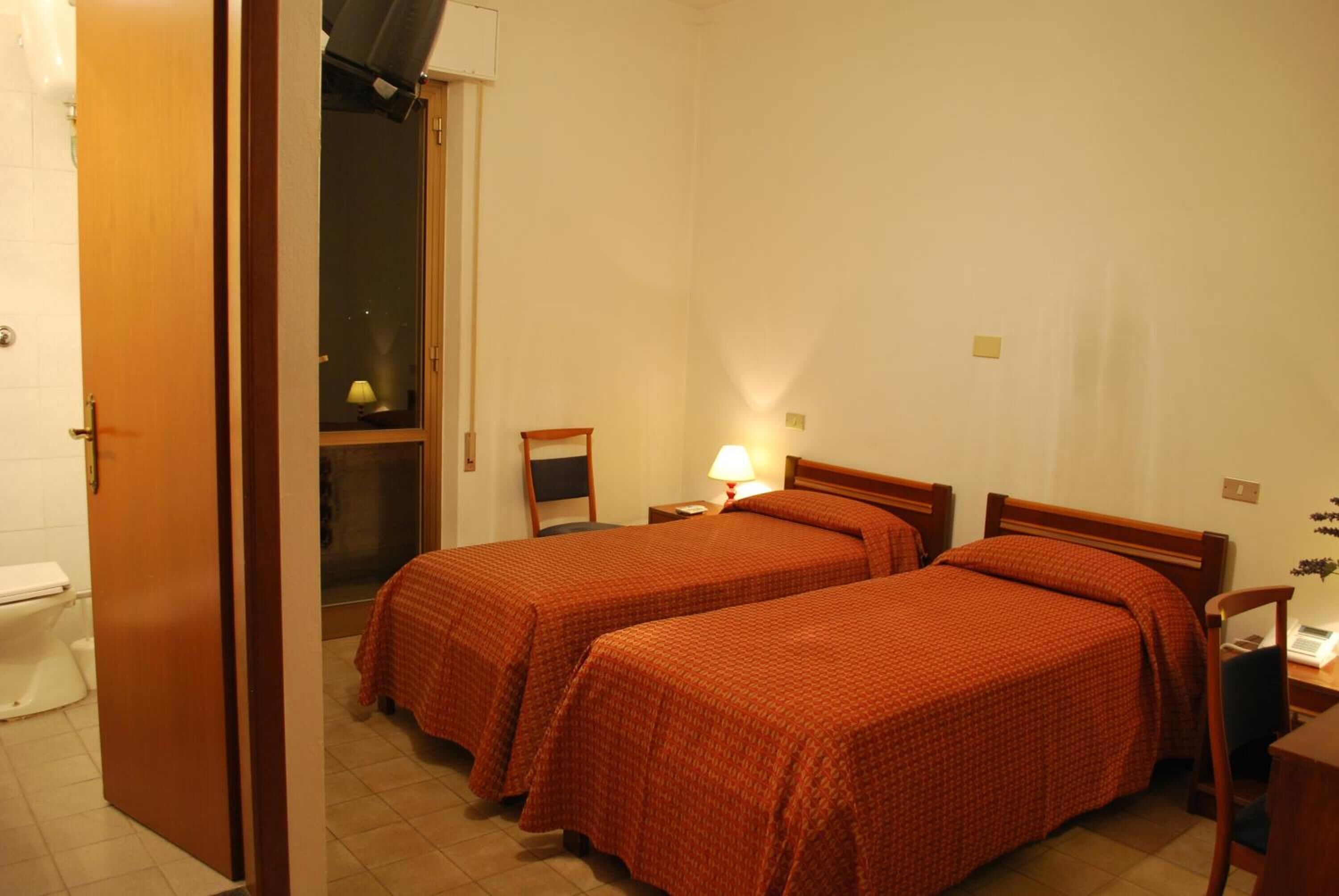 Una camera d'albergo con due letti, una porta di legno, un tavolino con una lampada e una finestra.