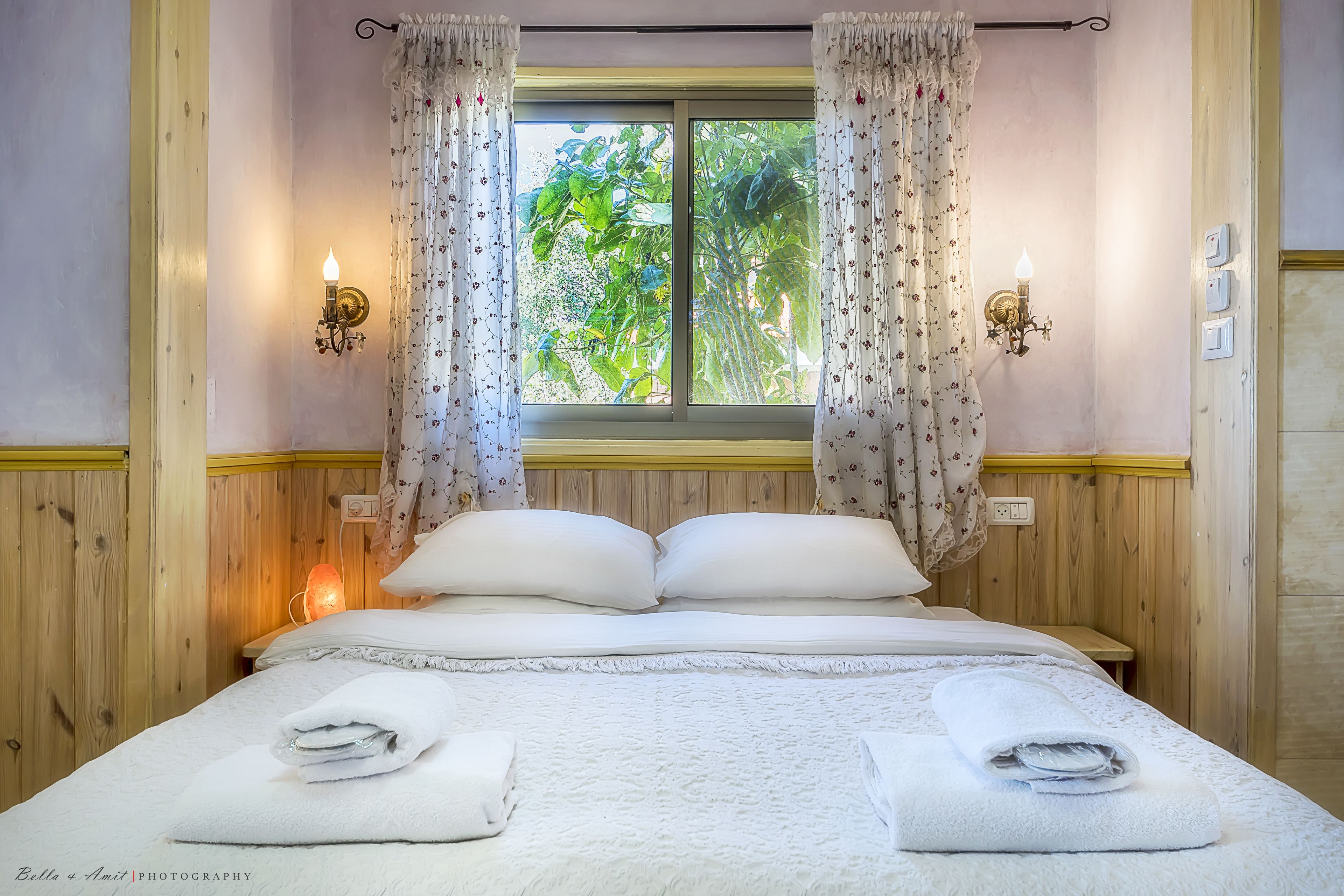 Una cama bien tendida con sábanas y almohadas blancas, una ventana con cortinas y luces fijadas a la pared.