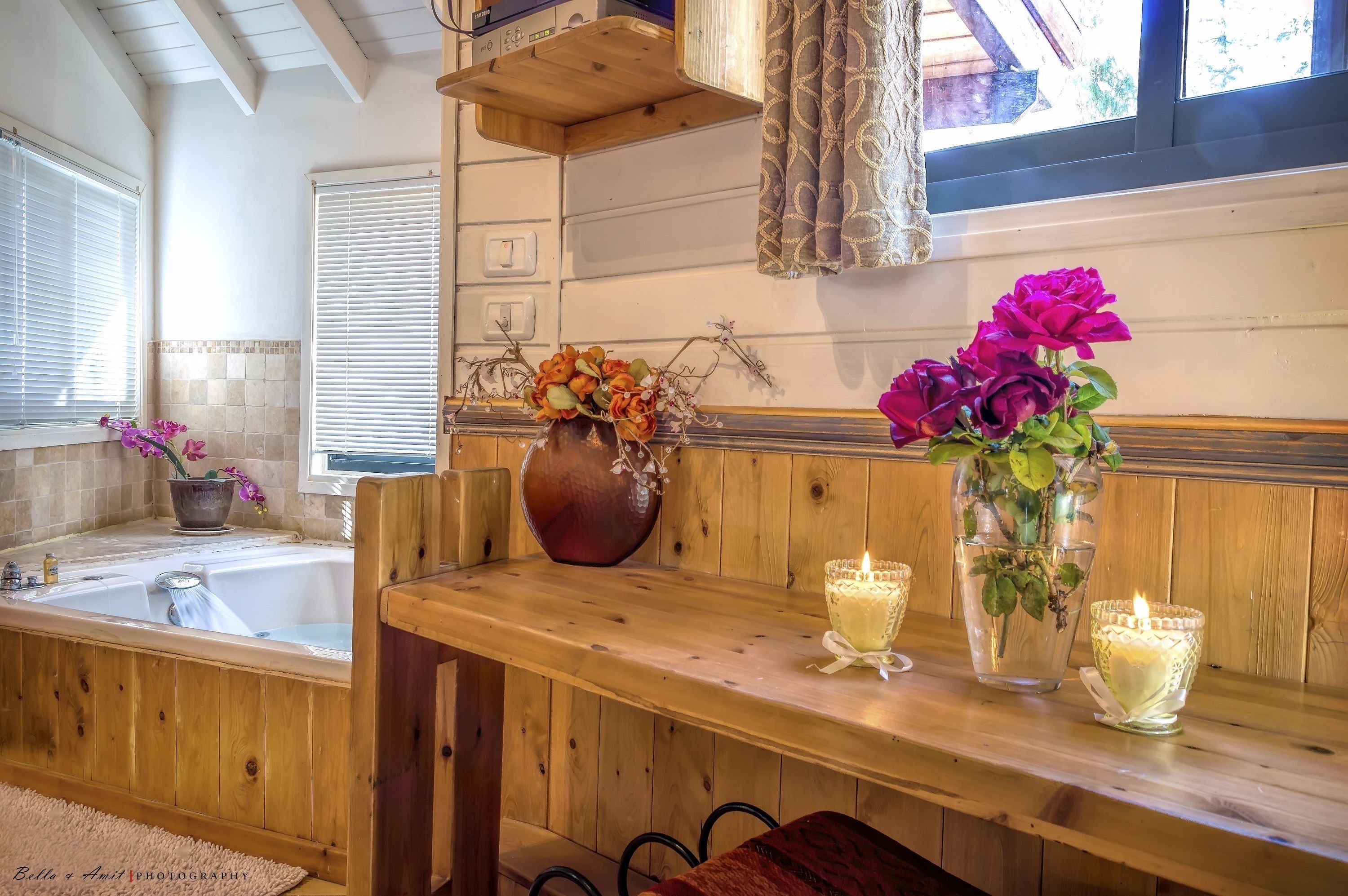 A wooden bench with a vase of flowers and candles, a bathtub, and a window with blinds.