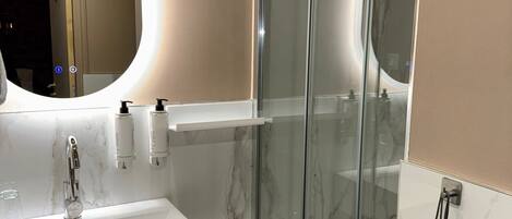 Un baño moderno con cabina de ducha de vidrio, un lavamanos blanco con espejo arriba y paredes de mármol.