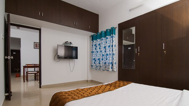 A bedroom with a bed, a TV mounted on the wall, a wardrobe, and a door leading to another room. - Phoenix Hospitality Services (Chennai)