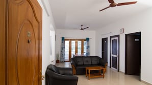 A spacious living area with a wooden door, a ceiling fan, a black leather sofa, a wooden coffee table, and a view through a glass door. - Phoenix Hospitality Services (Chennai)