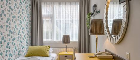 A hotel room with a bed, a desk with a telephone, a mirror, and a window with curtains.