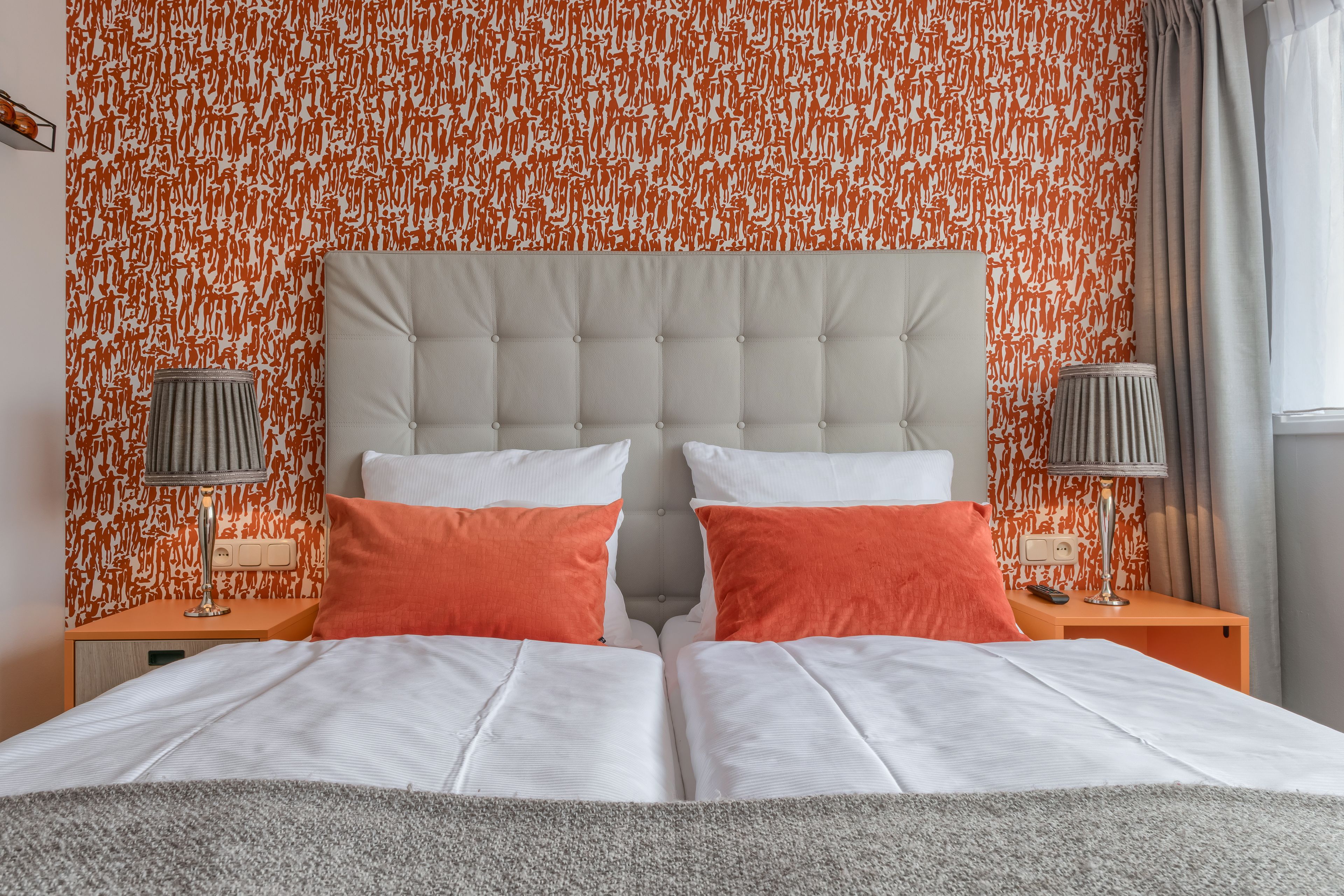 Ein Doppelbett mit weißer und orangefarbener Bettwäsche, einem gepolsterten Kopfteil und zwei Nachttischen mit Lampen.