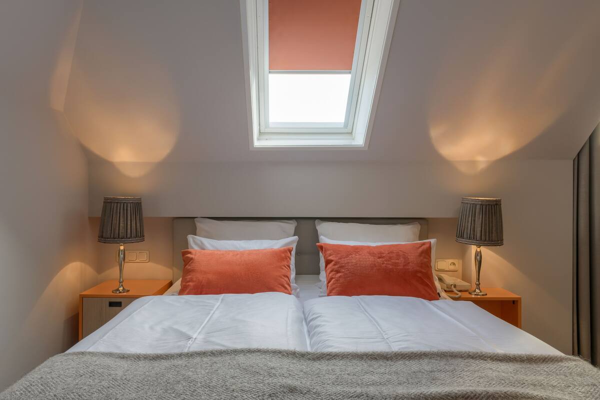 A bedroom with a skylight, two bedside tables with lamps, a bed with white bedding and orange pillows, and a telephone on one of the tables.
