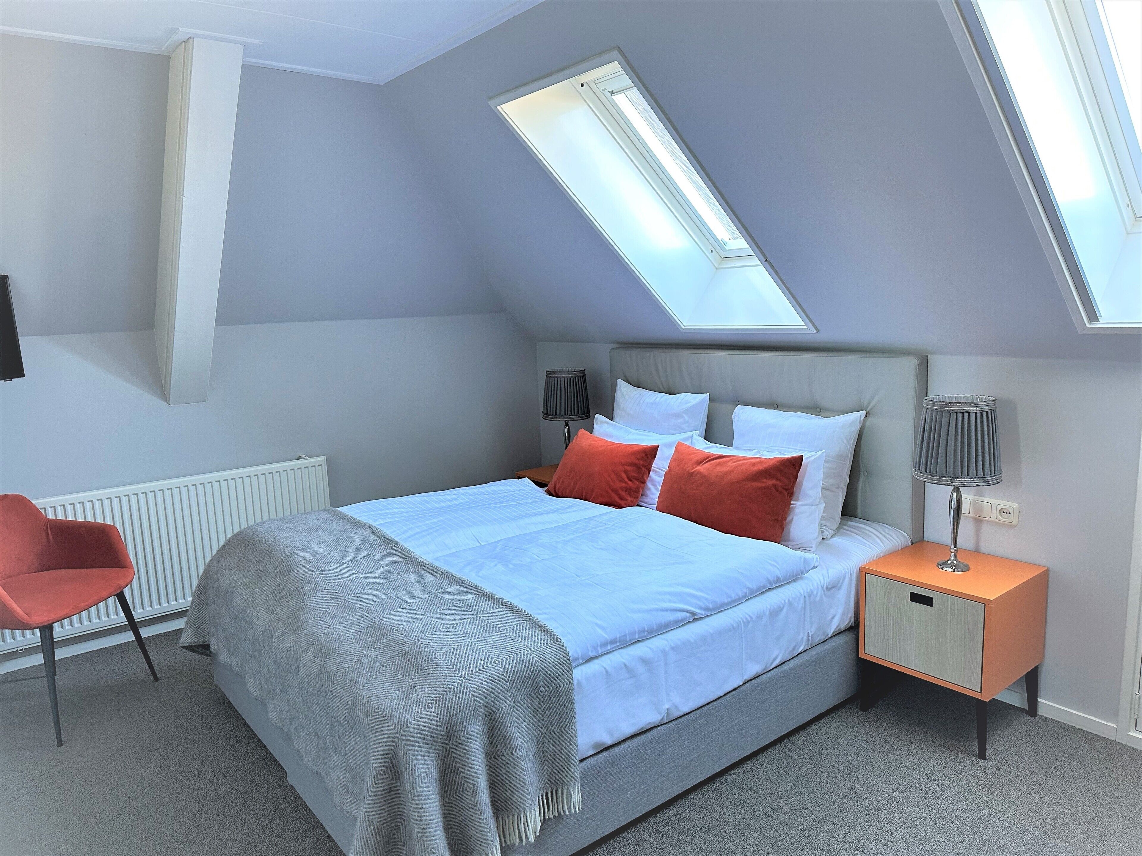 A bedroom with a skylight, a bed with white and orange pillows, a red chair, and a bedside table with a lamp.